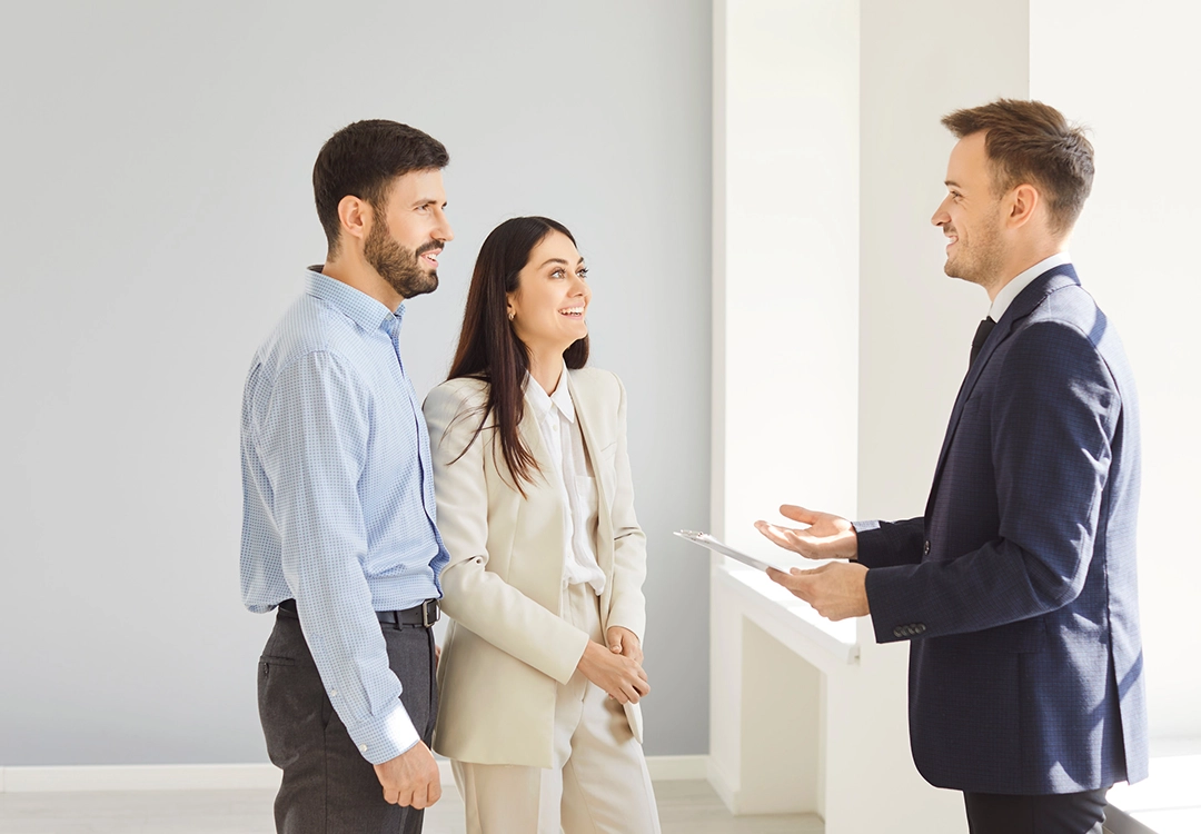 A professional meeting in a bright, modern setting, featuring a man and woman listening to a man in a suit discussing details.