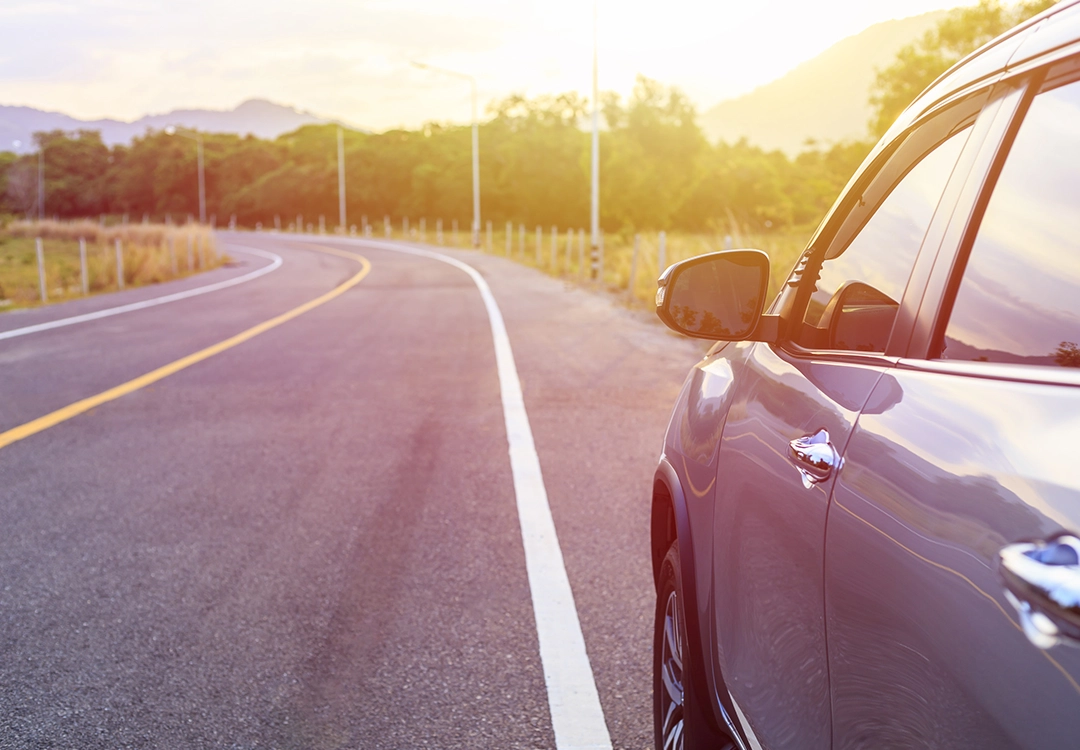 A close-up of a silver car parked on a winding road, surrounded by lush greenery and soft sunlight during golden hour.
